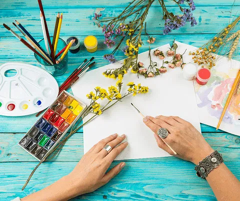 A person arranges yellow flowers beside paints and pencils on a blue table, highlighting creative work for a hyperlocal digital marketing agency in Pune. - IPSense.com